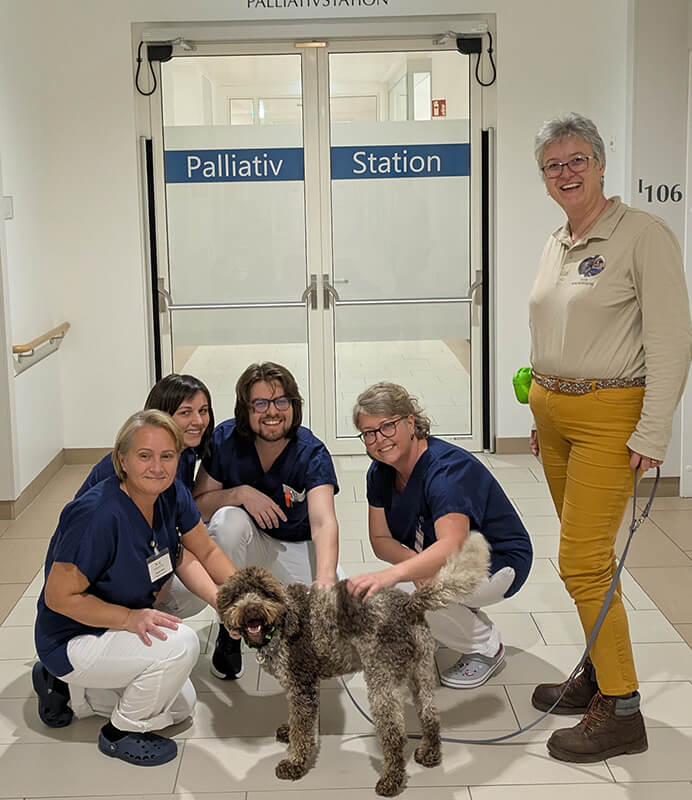 Therapiehund Osamu mit fünf lächelnden Teammitgliedern der Palliativstation im Franziskus Spital. Der Hund wird gestreichelt, im Hintergrund ist die beschriftete Glastür zur Station zu sehen.